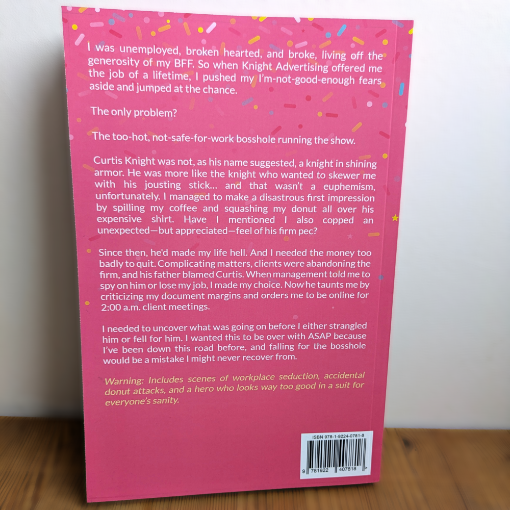 The back cover of a pink book titled 'Donut Girl and the Bosshole' resting on a wooden surface. The cover features a bright pink background with white text, colorful confetti accents, and a barcode at the bottom right.