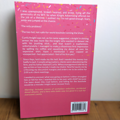 The back cover of a pink book titled 'Donut Girl and the Bosshole' resting on a wooden surface. The cover features a bright pink background with white text, colorful confetti accents, and a barcode at the bottom right.