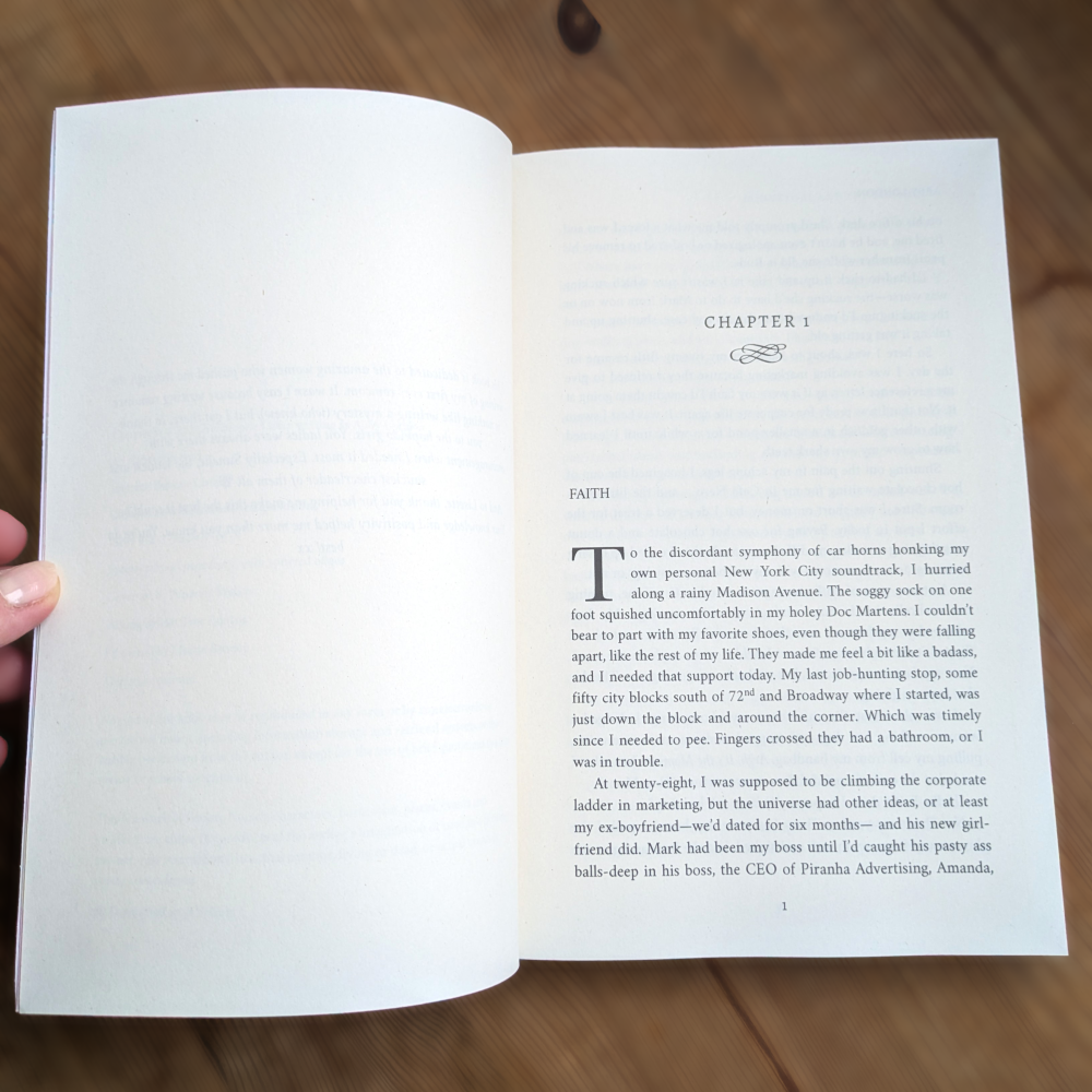 An open book showing the first chapter of Donut Girl and the Bosshole, held by a hand. The page has black text on warm off-white paper with a decorative chapter title 'FAITH' at the top.