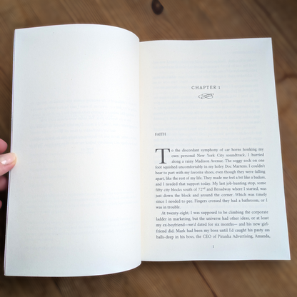 An open book showing the first chapter of Donut Girl and the Bosshole, held by a hand. The page has black text on warm off-white paper with a decorative chapter title 'FAITH' at the top.