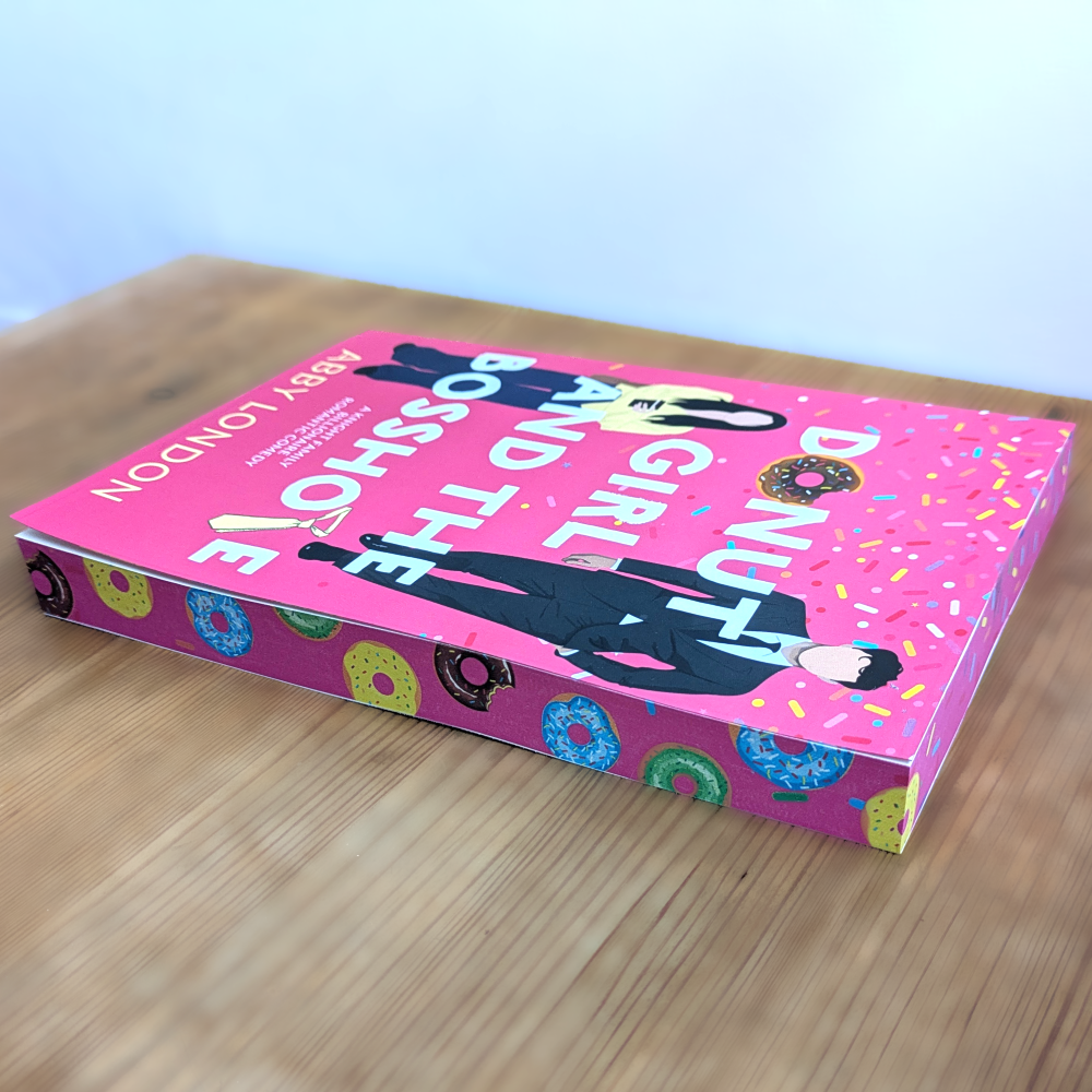A hardcover book titled 'Donut Girl and the Bosshole' by Abby London, featuring a bright pink cover with cartoon illustrations of a person in a black suit and colorful donuts, resting on a light wooden tabletop.