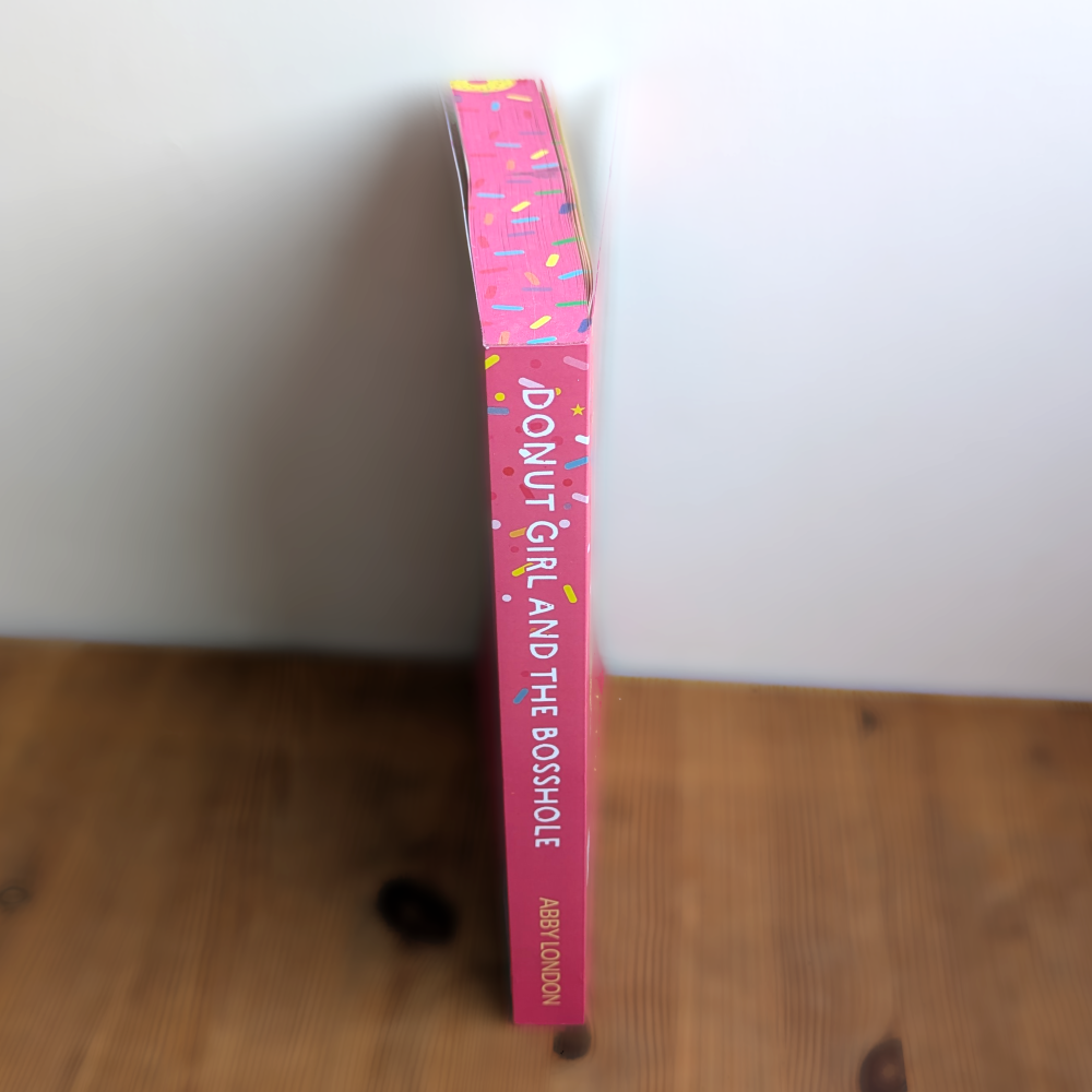 A pink hardcover book titled 'Donut Girl and the Bosshole' by Abby London, standing upright on a wooden surface against a plain white wall.