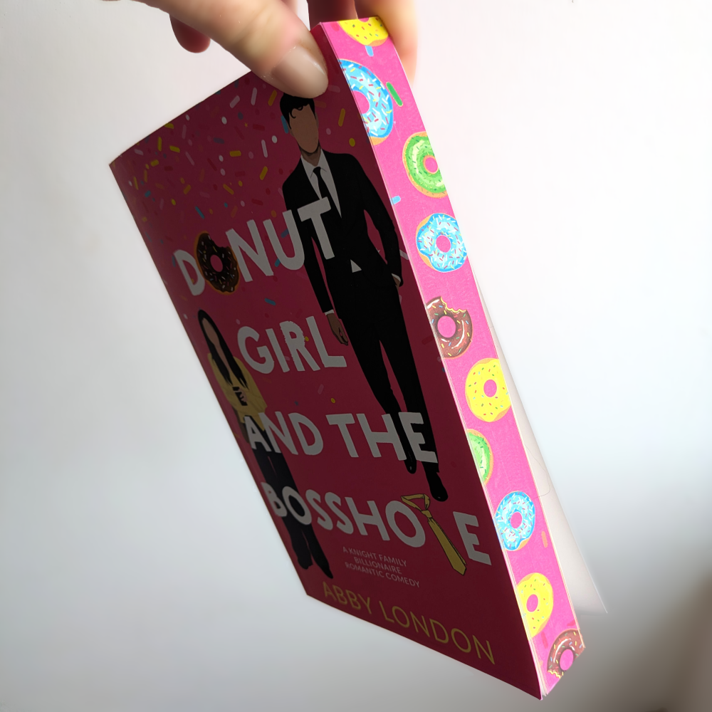 A hand holds a hardcover book titled "Donut Girl and the Bosshole" by Abby London. The book cover is dark pink with white text and an illustration of a man in a suit, while the spine features a bright pink background decorated with colorful donut illustrations.