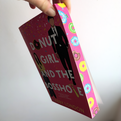 A hand holds a hardcover book titled "Donut Girl and the Bosshole" by Abby London. The book cover is dark pink with white text and an illustration of a man in a suit, while the spine features a bright pink background decorated with colorful donut illustrations.